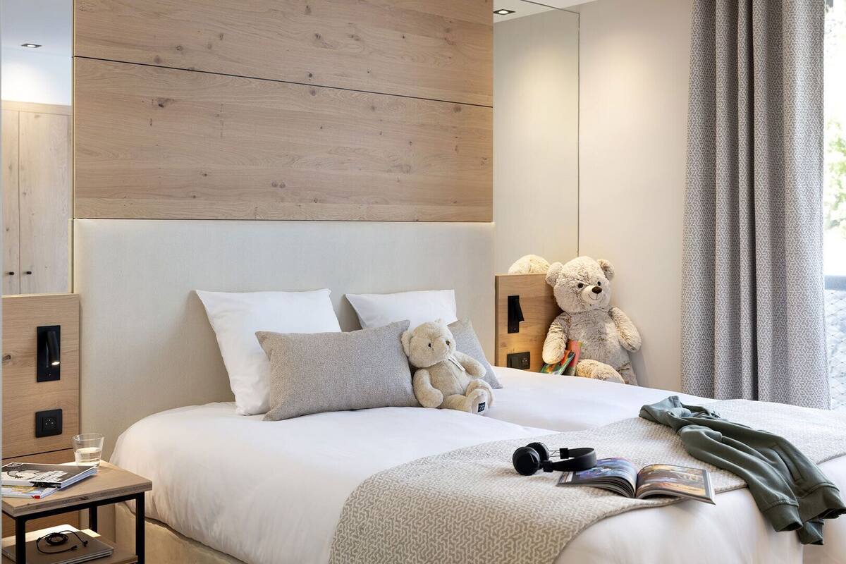 Un dormitorio moderno con cabecera de madera, cama con ropa blanca, dos almohadas, un oso de peluche, una mesita de noche con un vaso de agua, un libro y una chaqueta.