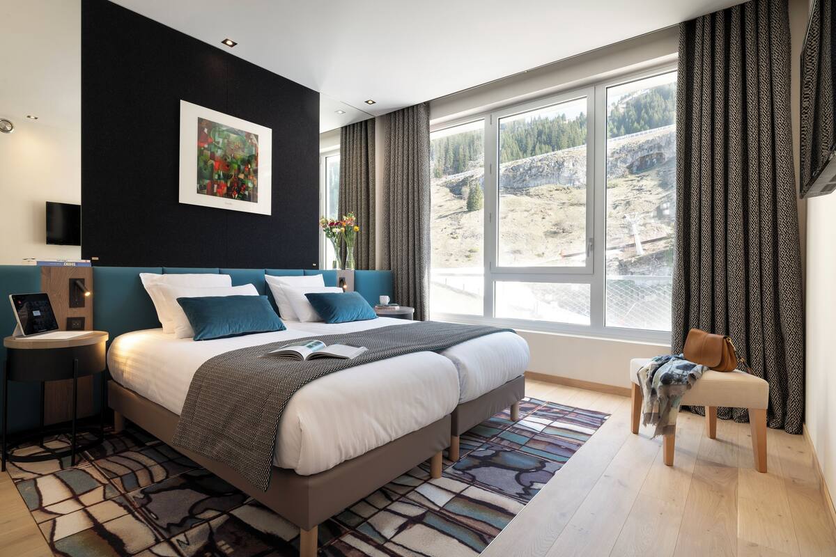 Un dormitorio moderno con una cama grande, un escritorio con una computadora portátil, vista a un paisaje montañoso y una ventana con cortinas estampadas.