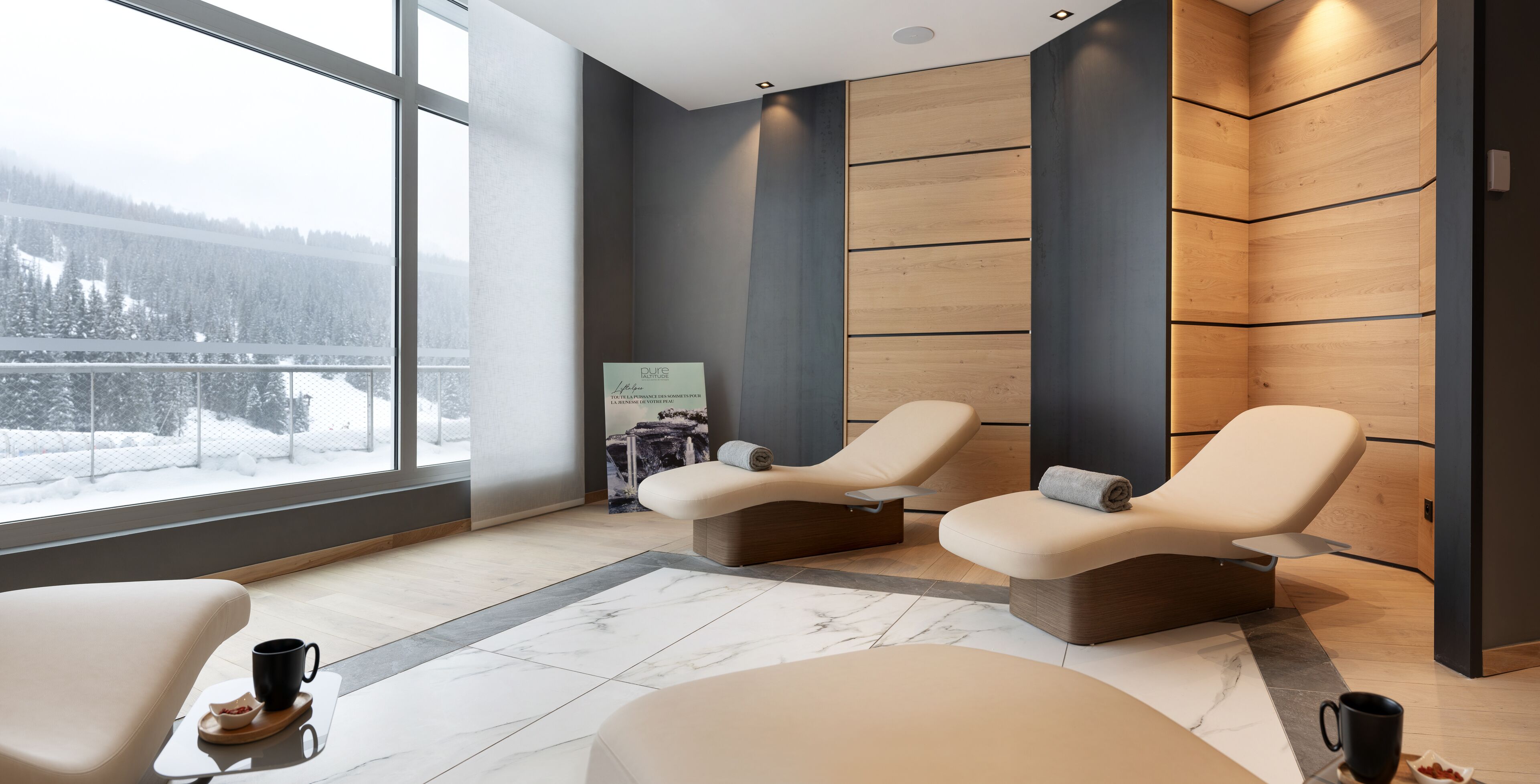 A modern spa room with two reclining chairs, a massage table, and a large window offering a snowy mountain view.