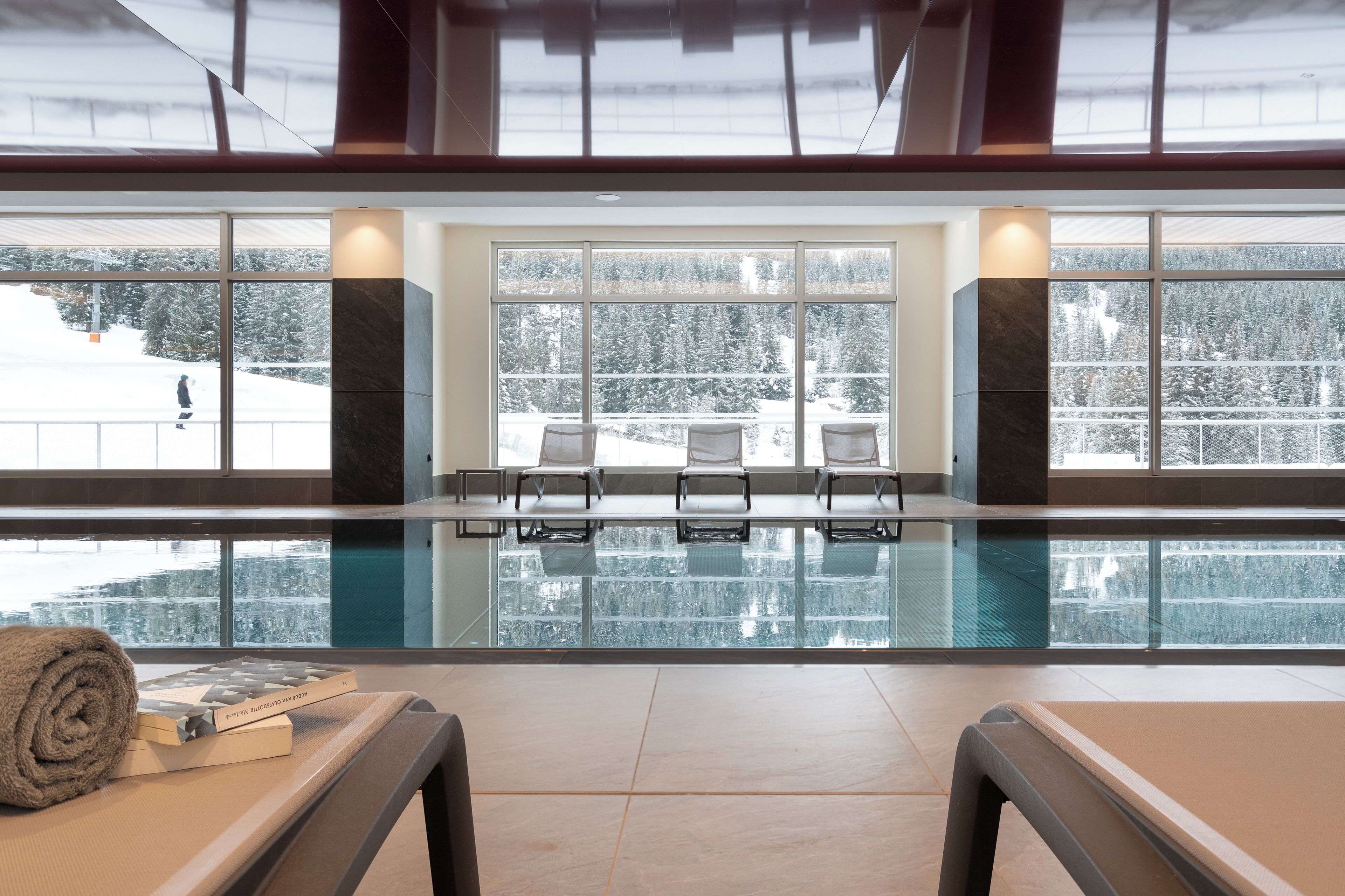 Ein moderner, überdachter Pool mit großen Glaswänden, Blick auf eine verschneite Landschaft und einigen Liegestühlen.