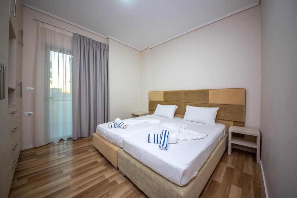 Een hotelkamer met twee bedden, een houten vloer, een groot raam met gordijnen en een nachtkastje.