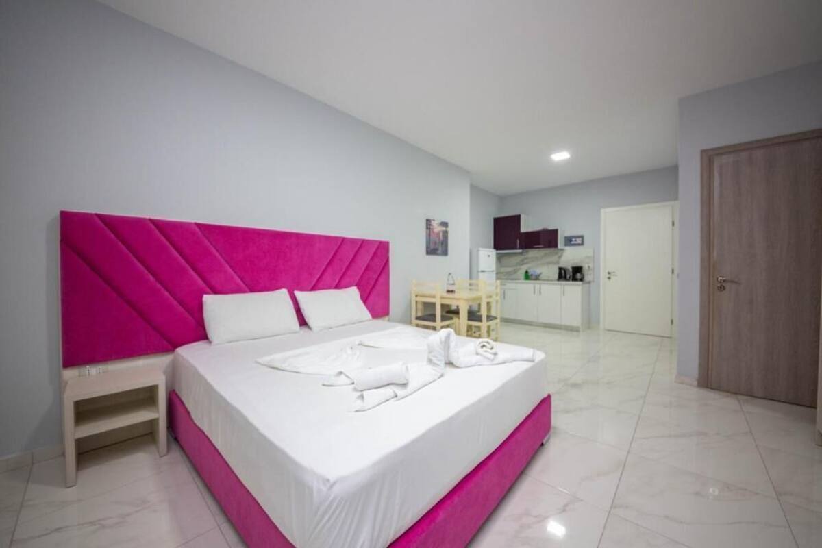 Una habitación de hotel moderna con una cama grande, una mesita de noche pequeña y una cocina americana al fondo.