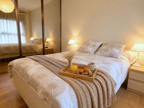 Un lit bien fait, avec un plateau contenant des articles pour le petit-déjeuner, un miroir qui reflète la pièce et une lampe de chevet.
