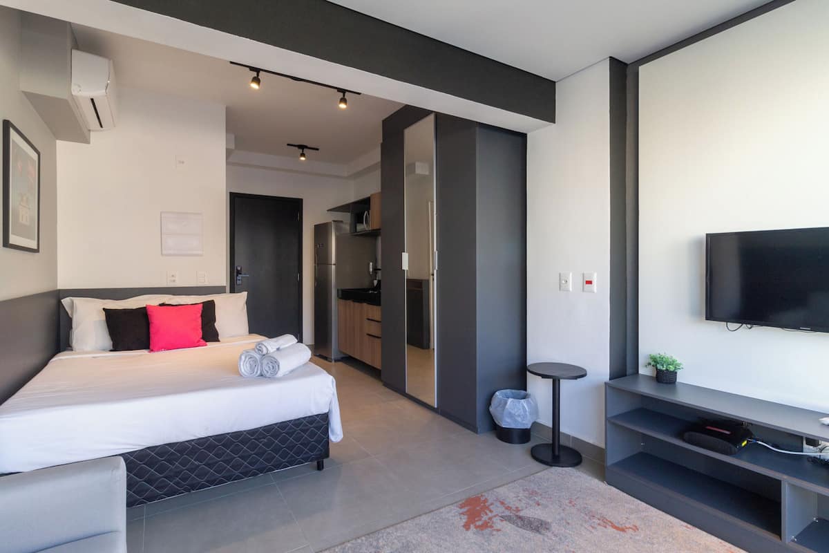 A modern hotel room with a bed, a TV, a small kitchen area, and a bathroom.