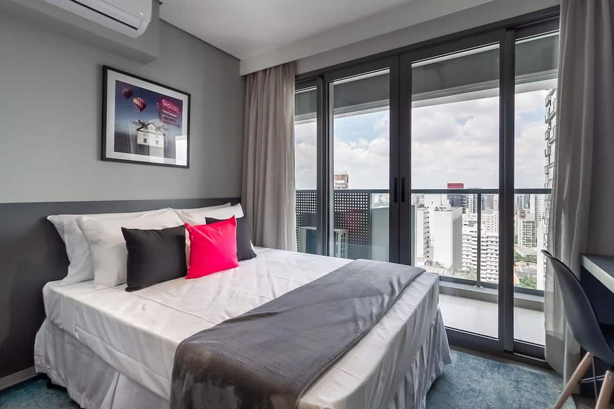 A hotel room with a large bed, a desk, and a view of the city through floor-to-ceiling windows.