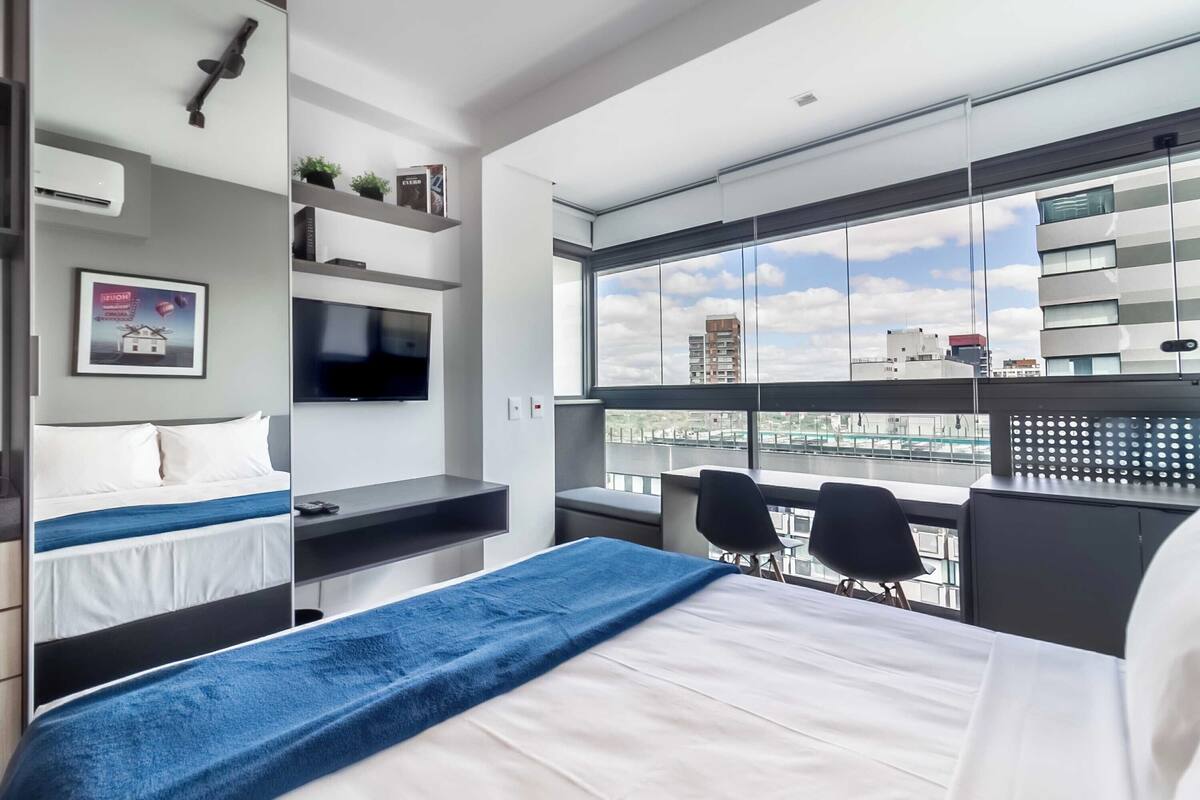 A modern hotel room with a large bed, a TV, a desk, and a view of the city through large windows.
