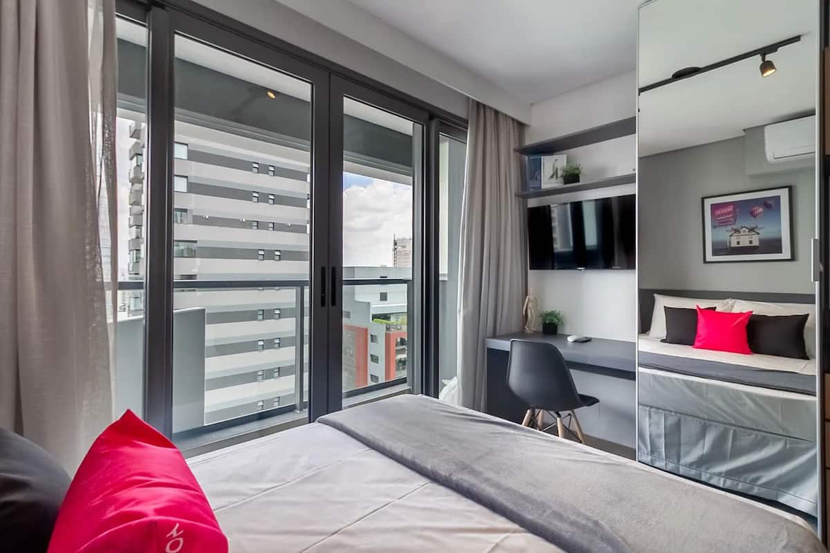 A modern hotel room with a large sliding glass door to a balcony, a bed with a grey and white duvet, a desk with a chair, and a mounted TV.