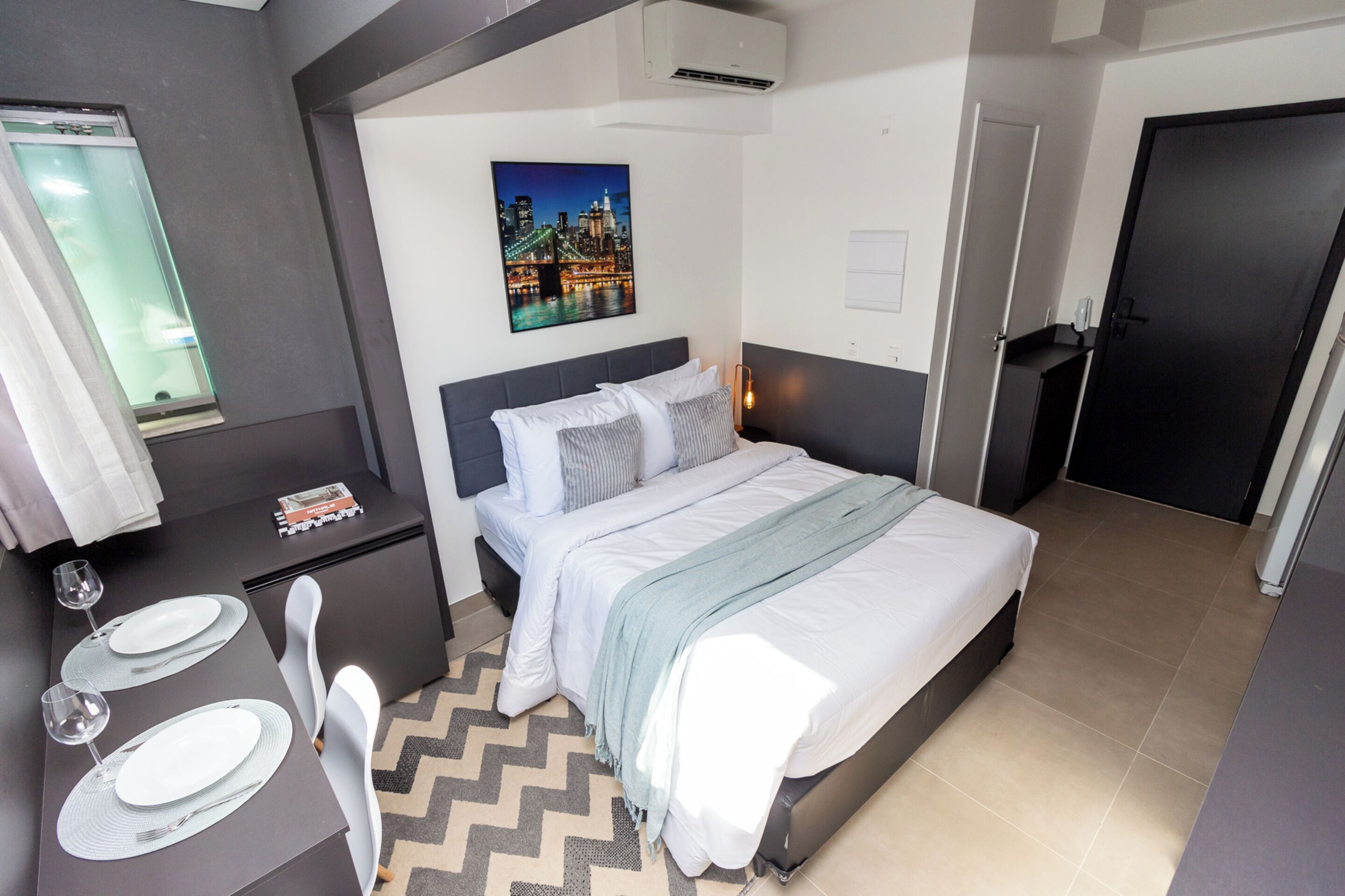 A modern hotel room with a bed, bedside table, and a cityscape painting on the wall.