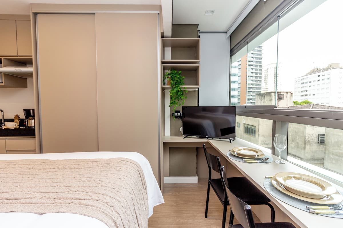 A modern hotel room with a bed, a desk with a chair, a plant, and a city view through the window.