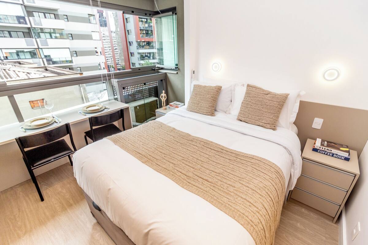 A hotel room with a bed, a desk with a chair, a small table with a wine glass, and a view of the city through a window.