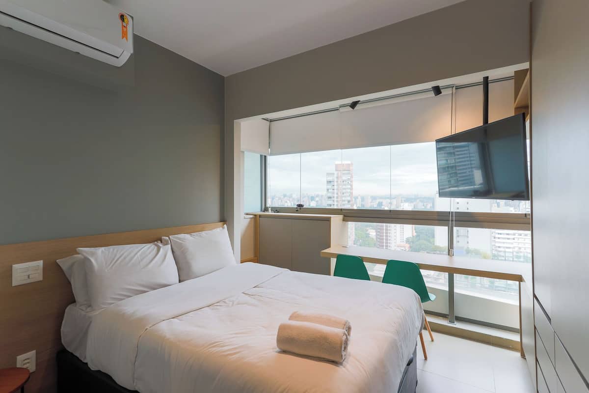 A modern hotel room with a large bed, a TV on a balcony, and a city view.