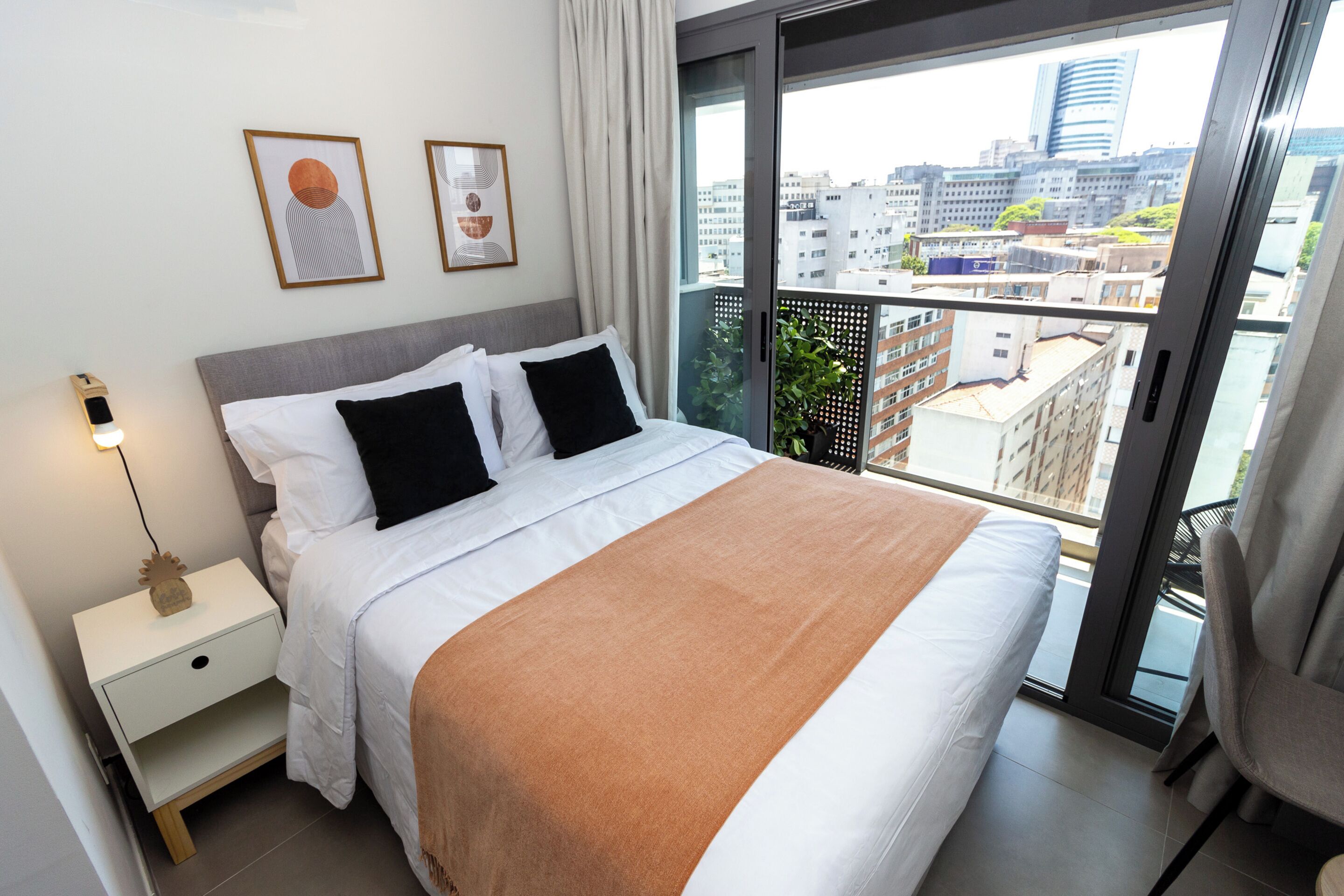 A modern hotel room with a large bed, a nightstand, a lamp, and a view of the city through a balcony.