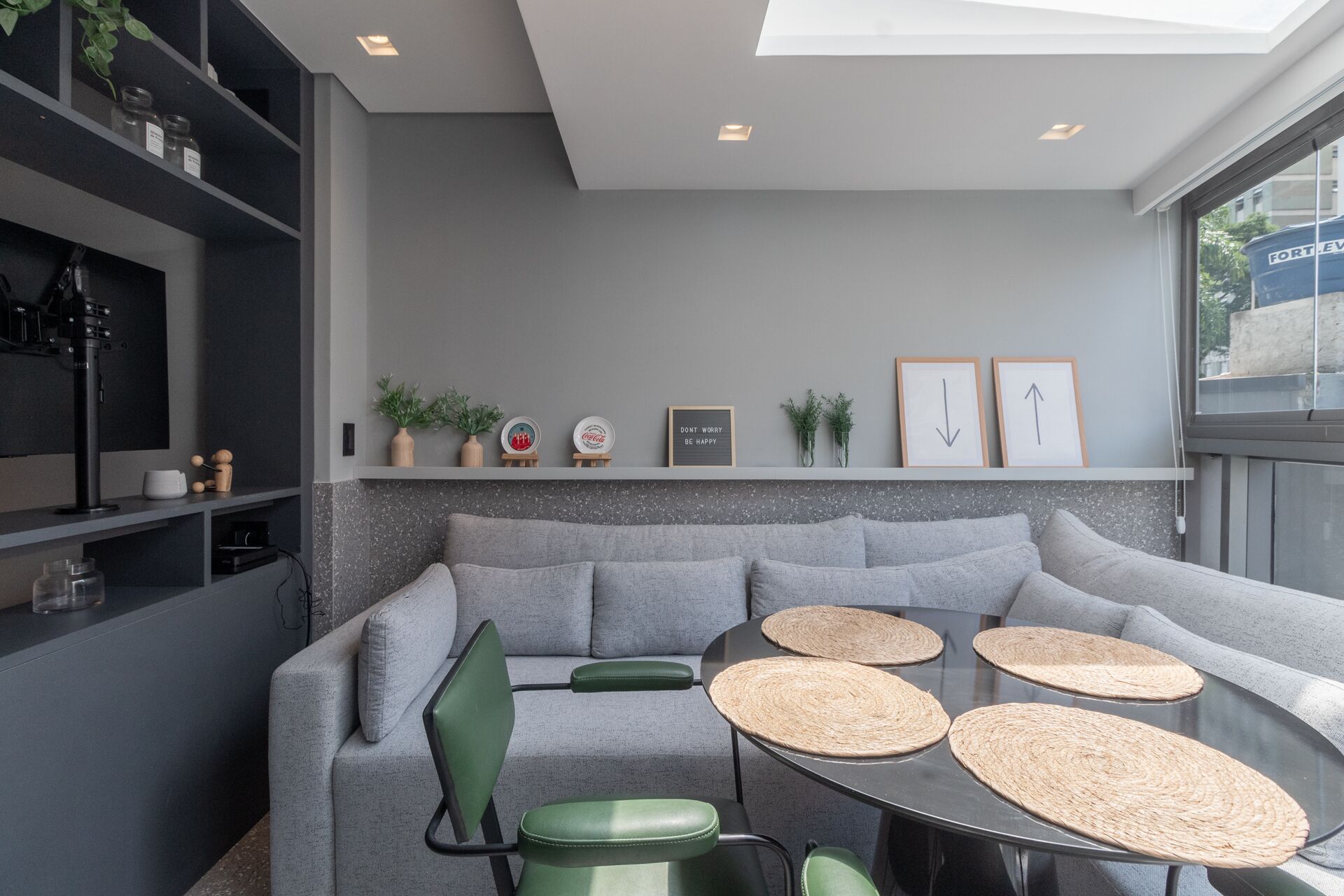 Un salon moderne avec un canapé gris, des fauteuils verts, une table ronde ornée de sets de table en fibres naturelles et une étagère contenant des objets décoratifs ainsi qu'un panneau indiquant : « N'AYEZ PAS D'INQUIÉTUDE, SOYEZ HEUREUX ».