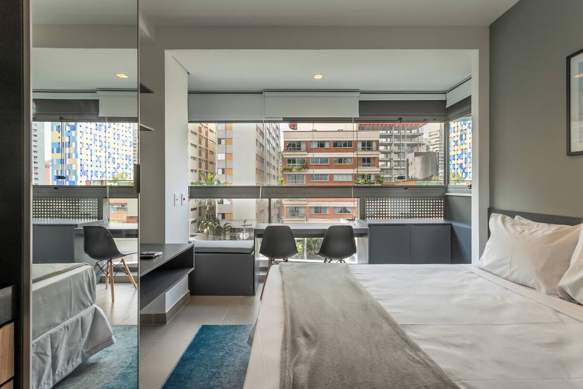 A modern hotel room with a large bed, a desk, two chairs, and a view of high-rise buildings.