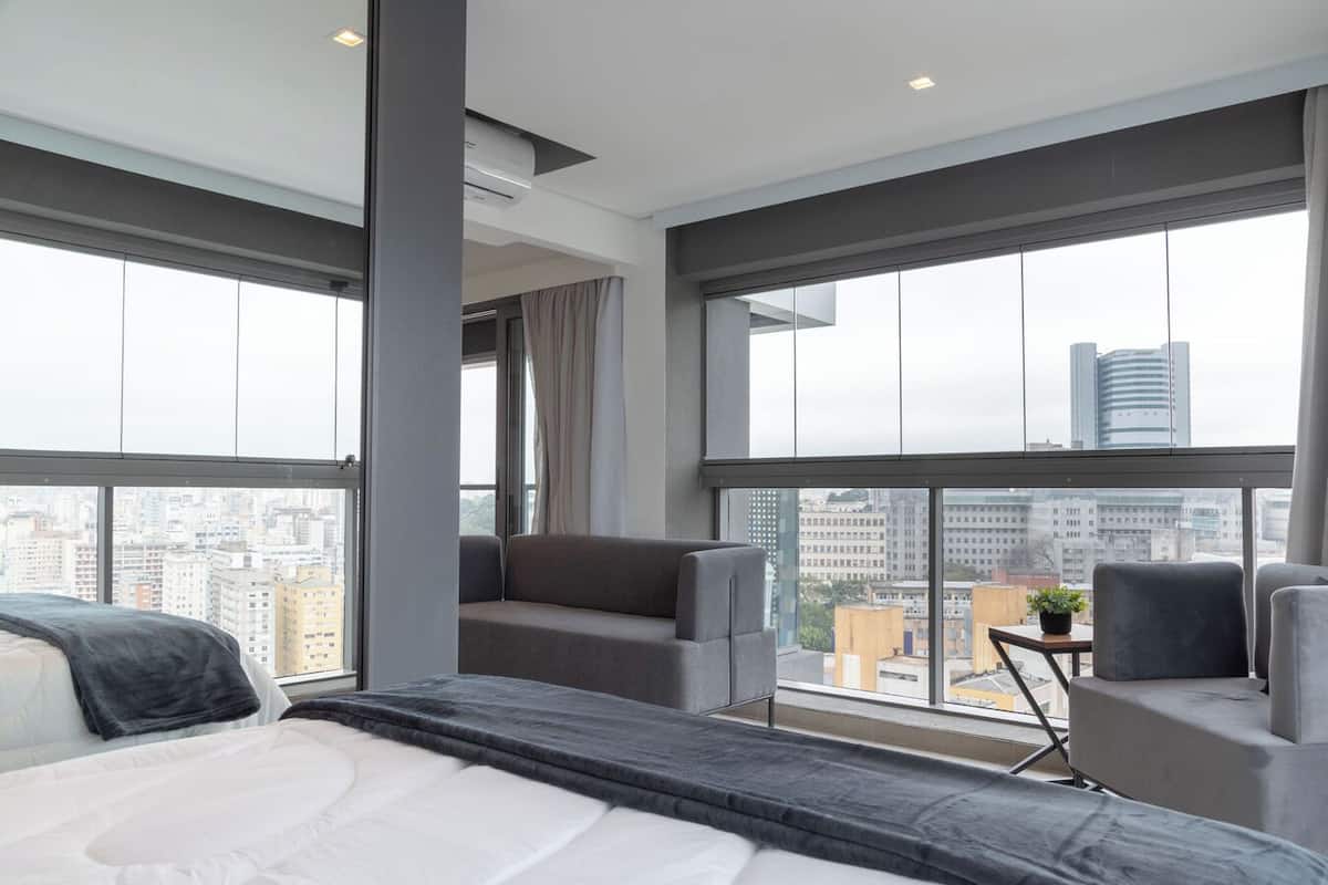 A modern hotel room with a large bed, a seating area with a small table, and a city view through large windows.