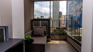 A small balcony with a sofa, a potted plant, and a view of the cityscape through large windows.