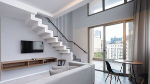 A modern living room with a staircase, a flat-screen TV, a sofa, a dining table, and a view of the city through large windows.