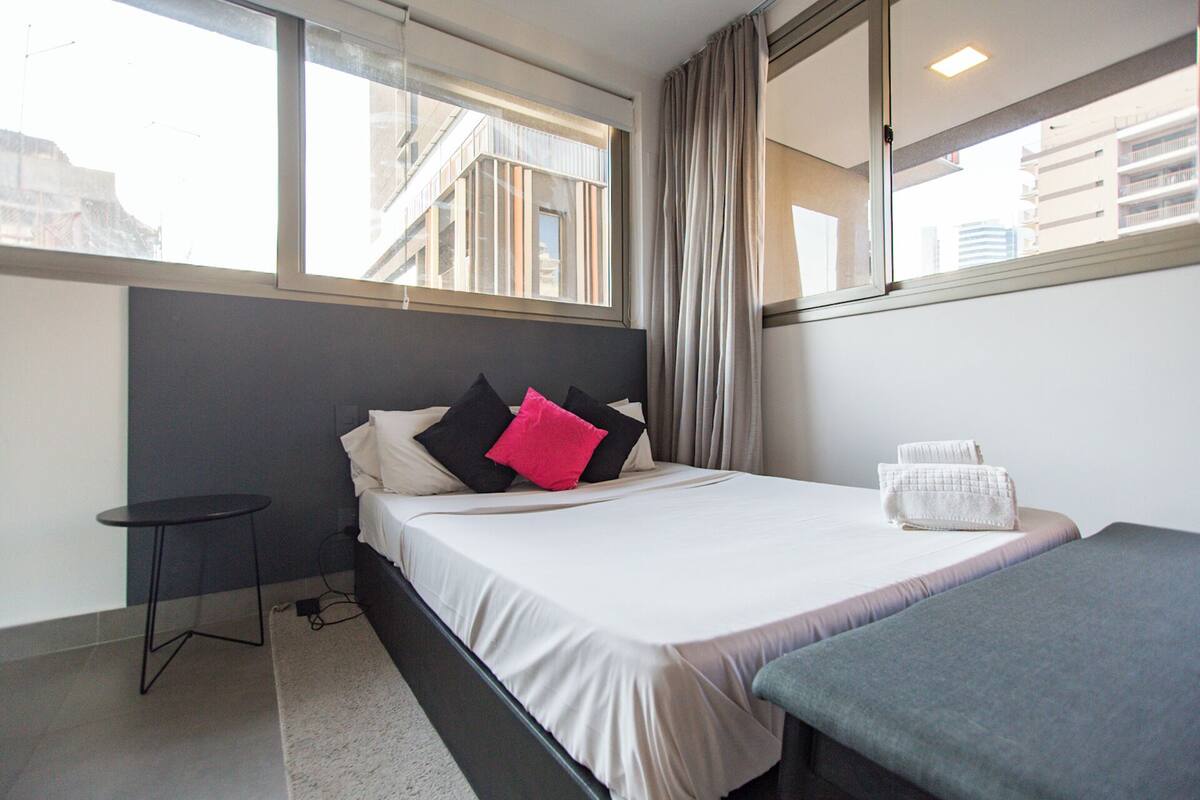 Ein Hotelzimmer mit Bett, kleinem Tisch, Nachttisch, Fenster und Blick auf Gebäude.
