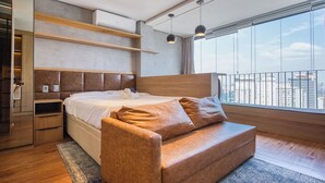 A modern bedroom with a bed, a wooden headboard, a built-in shelf, a window with a city view, and a balcony railing.