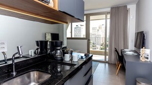A modern kitchen with a black countertop, stainless steel sink, and a coffee maker. There is a dining area with a table and chairs, and a view of the city through large windows.