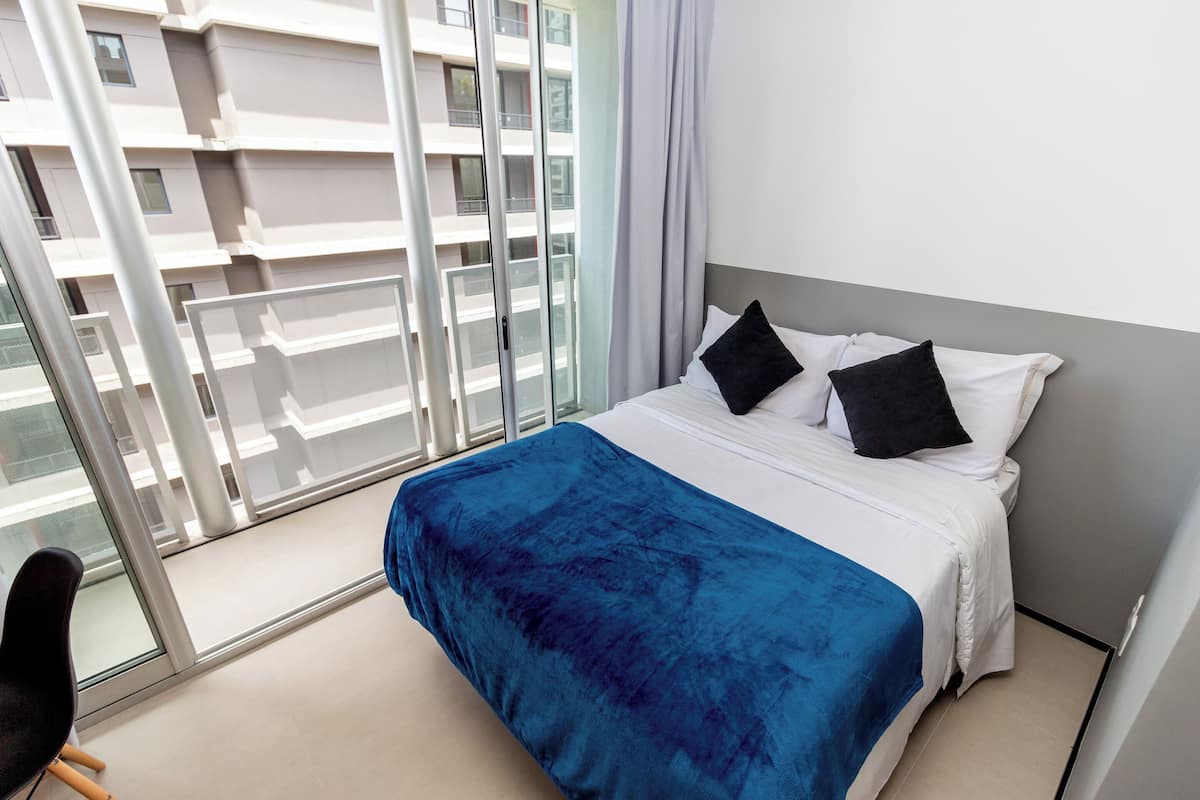 A modern hotel room with a large bed, a view of the city through floor-to-ceiling windows, and a balcony with a glass railing.