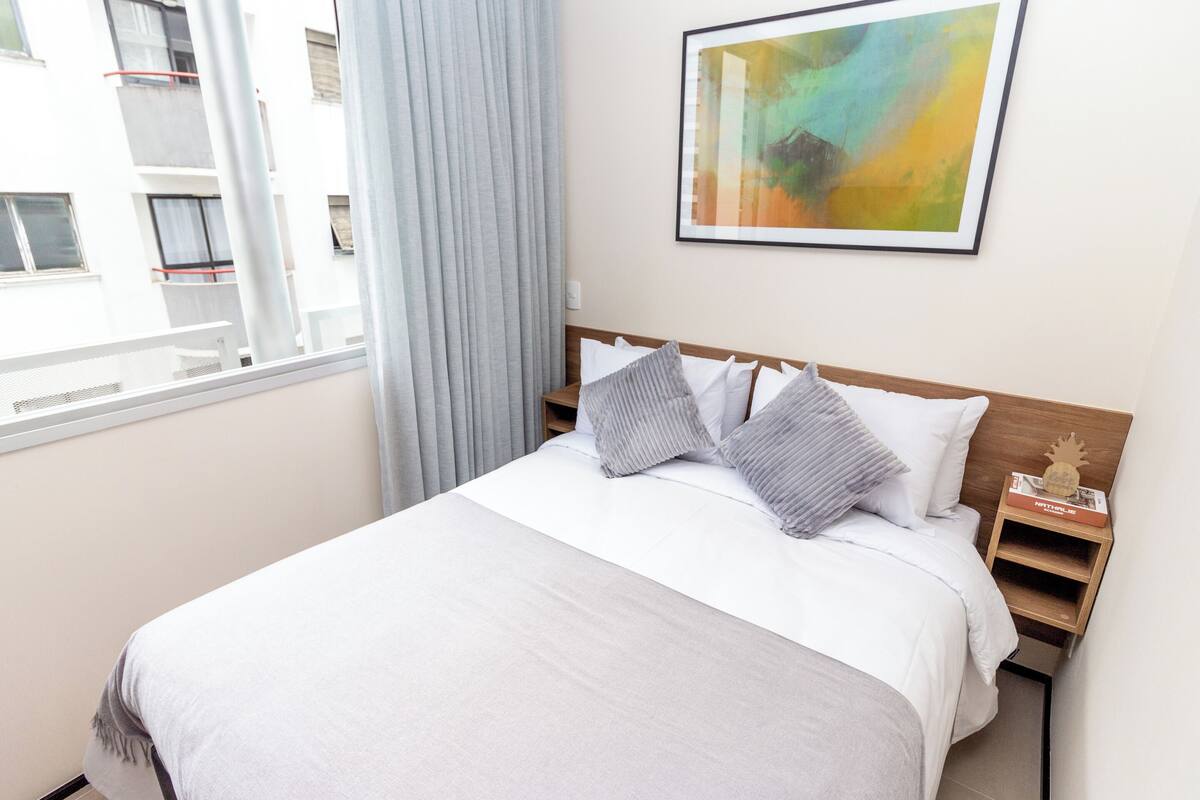 A neatly made bed with white bedding and two pillows, a wooden nightstand with a book and a pineapple decoration, and a colorful abstract painting on the wall.