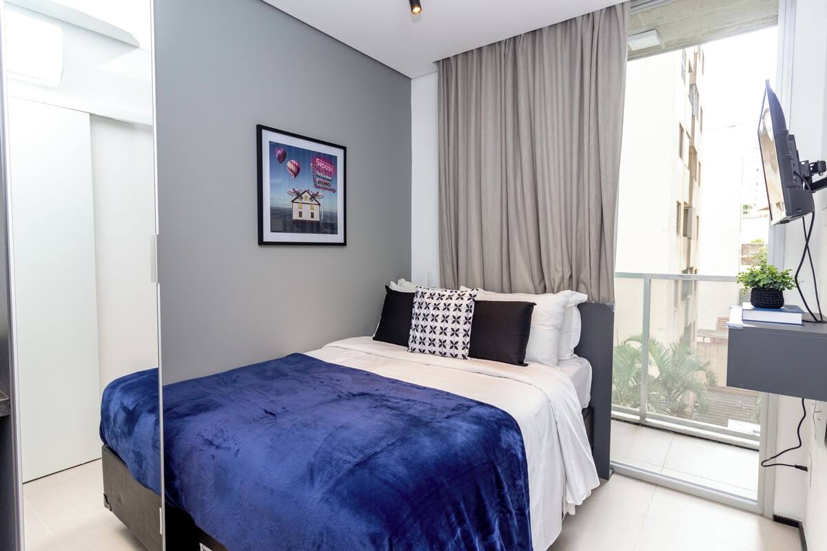 A modern bedroom with a large bed, a TV, and a balcony with a view of buildings.