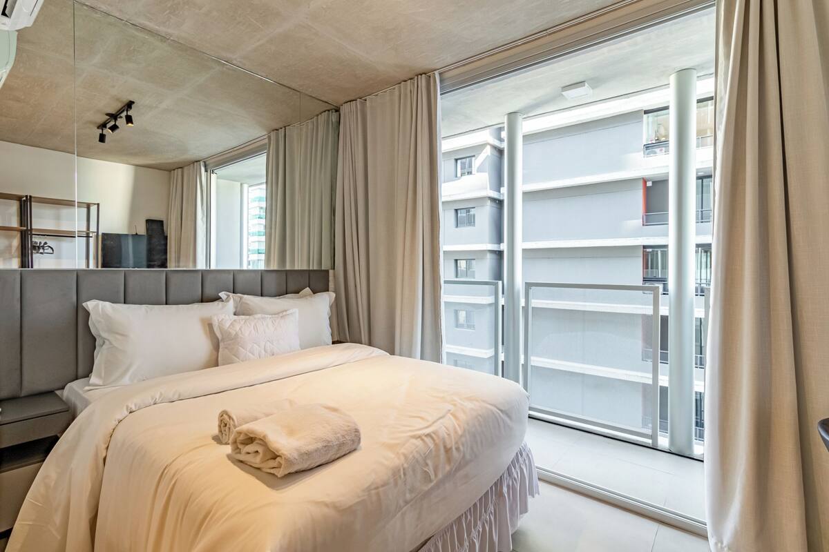 A modern hotel room with a large bed, a nightstand, a mirror, and a view of the building's exterior through a large window.