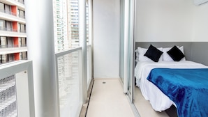 Un balcón estrecho con barandilla de metal, una cama con ropa blanca y almohadas negras, y una colcha azul.
