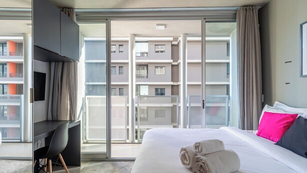 Habitación de hotel con cama, escritorio, silla y un amplio ventanal con vistas a otros edificios.