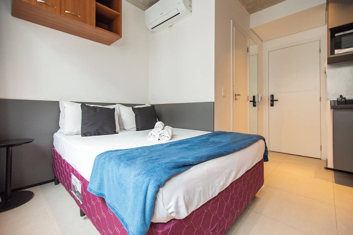 A hotel room with a bed, a small table, a wardrobe, and an air conditioning unit.