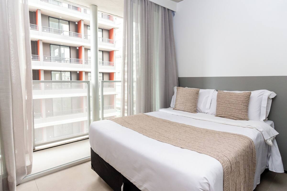 A hotel room with a large bed, a window with a view of a building, and a curtain.