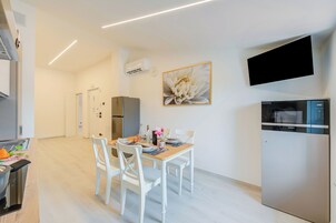 Una cocina moderna con zona de comedor, sillas blancas, una mesa con un florero y un televisor montado.