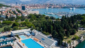 Paisagem costeira com um porto, uma piscina e uma fortificação histórica.