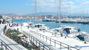 Marina com vários iates atracados, um passeio para pedestres e paisagem urbana ao fundo.