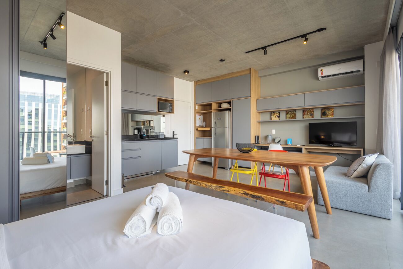 A modern hotel room with a bed, a wooden bench, a dining table, a kitchen area, and a living space with a TV.