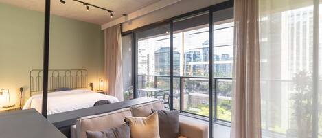 A modern hotel room with a large window, a bed, a sofa, and a desk.