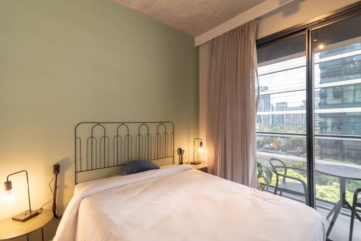 Habitación de hotel con cama, lámparas de noche, ventana con vistas a edificios y balcón con sillas.