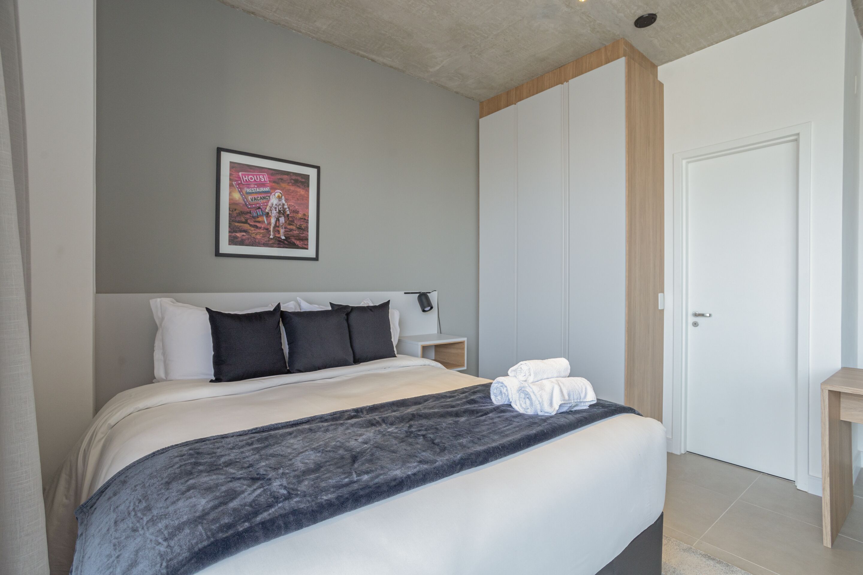 Quarto de hotel moderno com uma cama grande, um quadro na parede e uma porta de madeira.