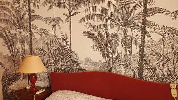 A room with a bed, a nightstand, a lamp, and a wall with a tropical mural.