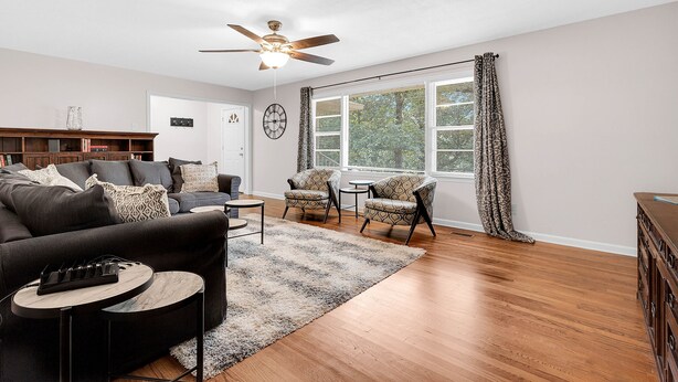 A modern living room with a dark sofa, a coffee table, patterned chairs, a ceiling fan, and a large window with curtains. - Fantastic for Extended Stays & Work Crews- 10 beds, 5 bedrooms, 2.5 baths (Warner Robins)