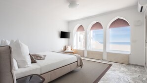 Een moderne hotelkamer met een bed, een klein tafeltje, een televisie en uitzicht op het strand door boogvormige ramen.