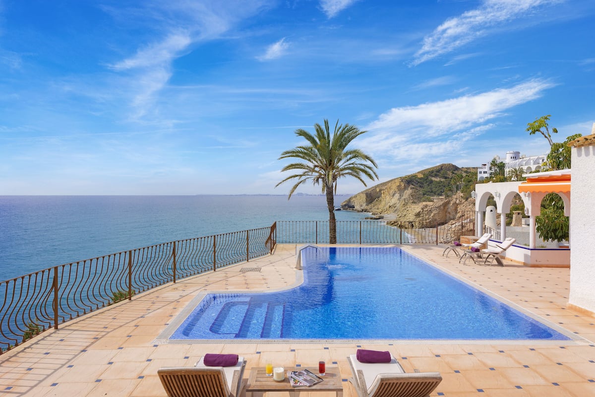 A pool with a view of the sea, a palm tree, and lounge chairs.