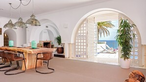 Een ruime woonkamer met een houten eettafel, lederen stoelen en hanglampen. Een grote potplant is zichtbaar, en een open boog leidt naar een balkon met uitzicht op zee.
