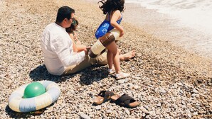Een stel zit op een kiezelstrand met een rotsachtige klif en palmbomen op de achtergrond.