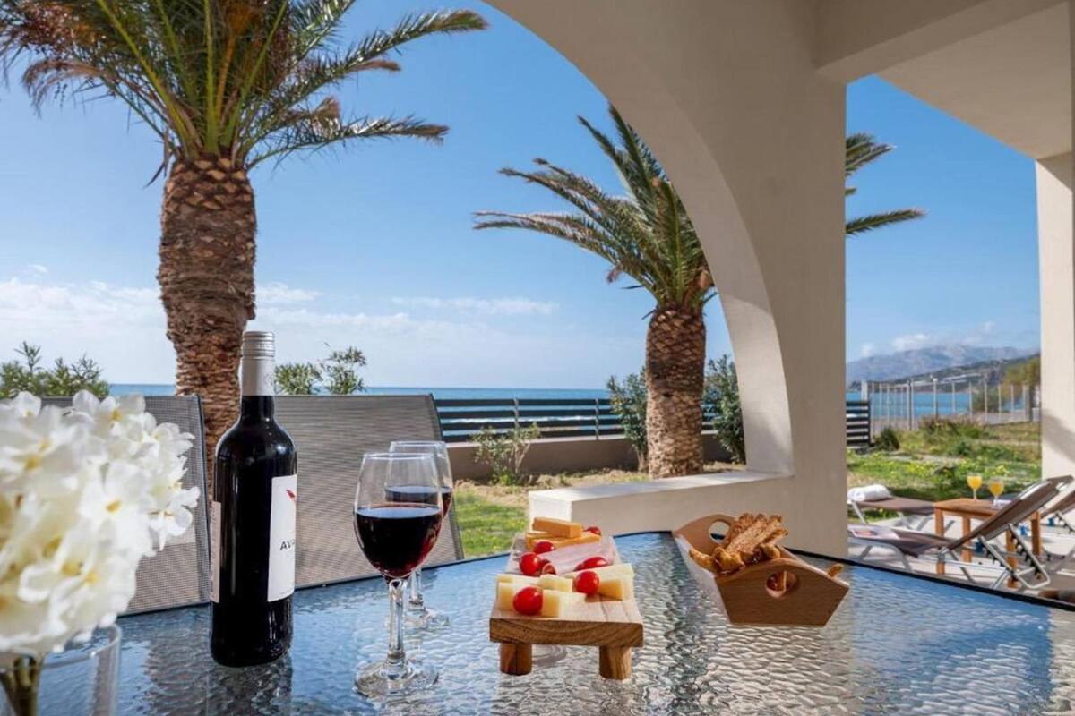 Uma mesa com uma garrafa de vinho, um copo de vinho tinto, uma tábua de madeira com queijo e tomate, e um vaso com flores brancas.