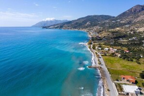 Vista aérea de uma cidade costeira com mar azul-turquesa, uma estrada estreita ao longo da costa e terreno montanhoso ao fundo.