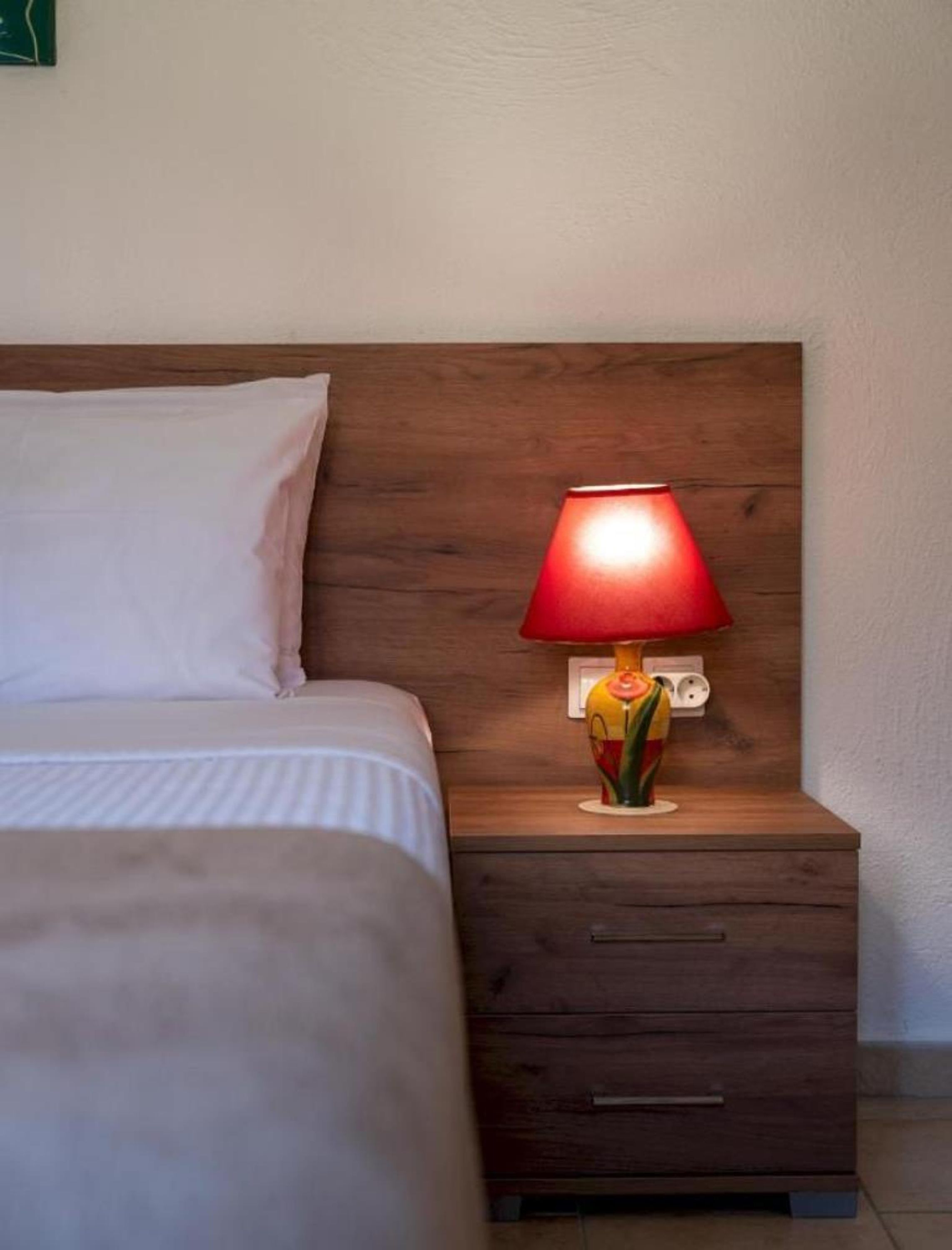 A neatly made bed with a wooden headboard, a bedside table with a red lamp, and a small drawer.
