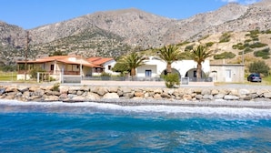 A coastal property with a stone wall, a white building with a red roof, and a mountain backdrop. - Atalanti Seaside Junior Suites (Viannos)
