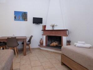 A room with a fireplace, a TV mounted on the wall, a dining table with chairs, and a bed. - Atalanti Seaside Junior Suites (Viannos)